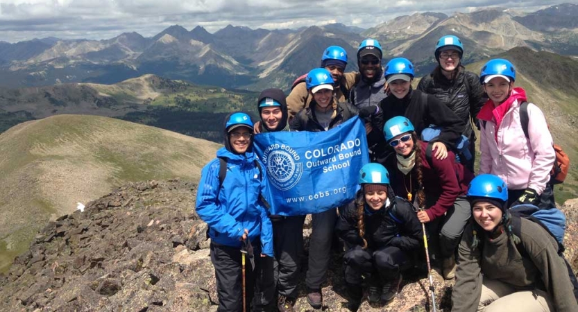 A group of students wearing helmets pose on top of a peak with a mountainous landscape in the background. 